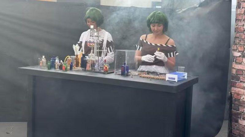 Two young women dressed as Oompa Loompas, in front of a makeshift chemistry set.