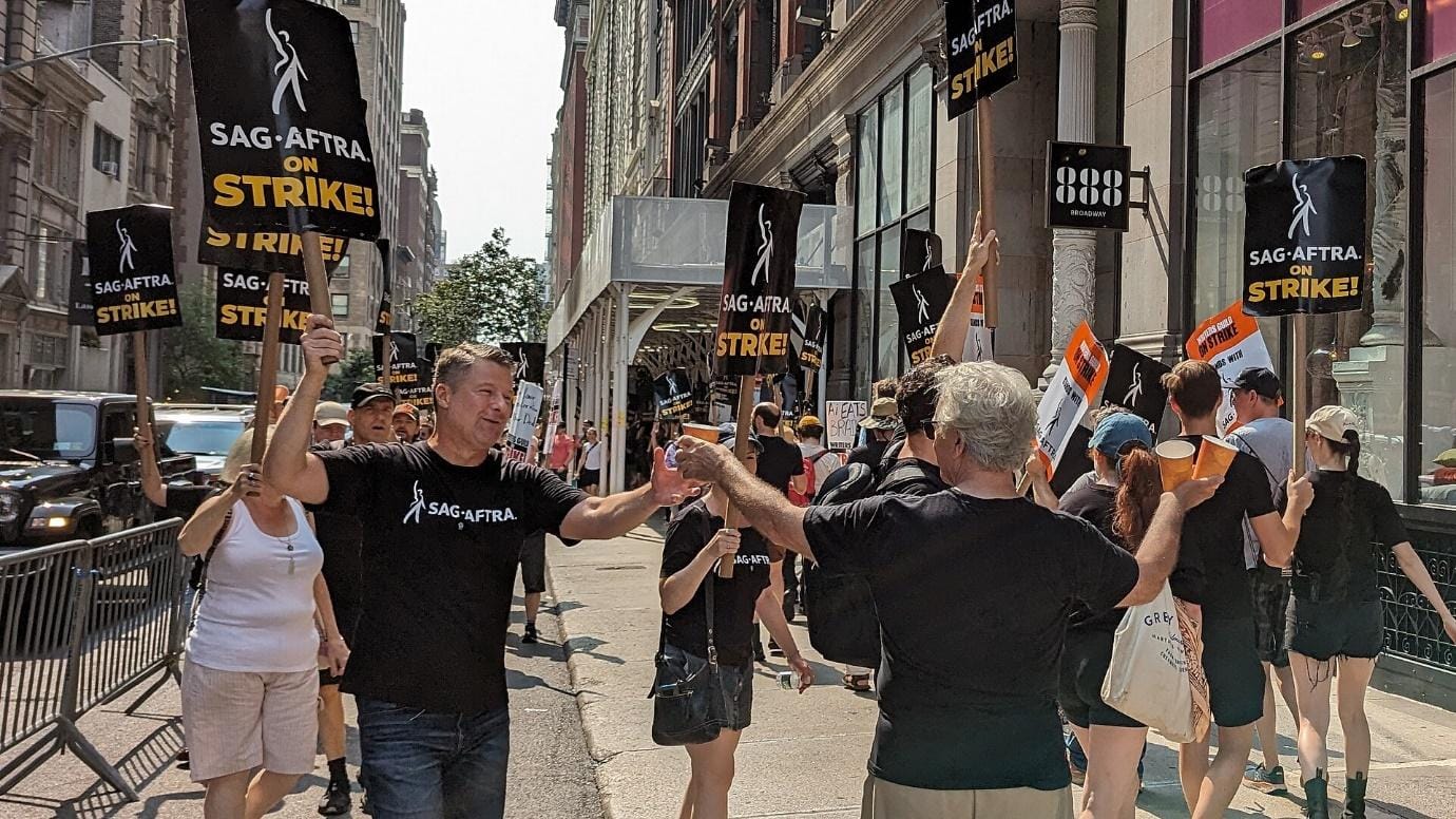 Workers in black SAG-AFTRA t-shirts picket outside a theatre holding 'SAG-AFTRA on strike' signs.
